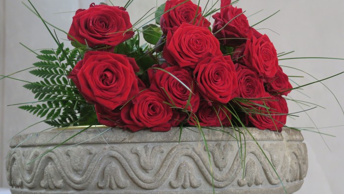 Man sieht den oberen Teil des Taufsteines in der Johanneskirche, er ist aus Sandstein und verziert, darauf liegen dekorativ einige rote Rosen.