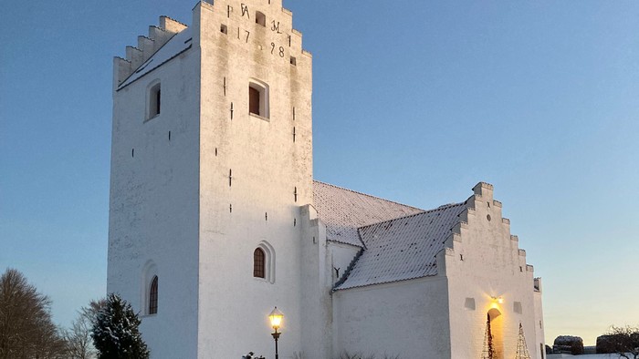 En snede hvid kirke med tårn og en lystet indgang i snedæknede omgivelser