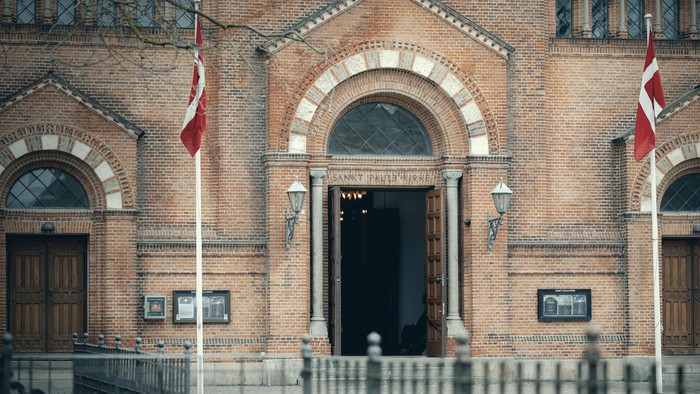 Bygningen har to danske flag og en central indgang med bueformet vindue.