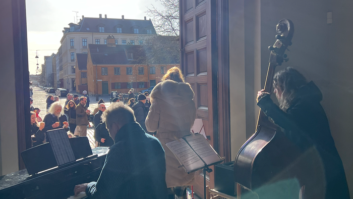 Musikere spiller på instrumenter indendørs, mens en folkemængde samles udendørs i et solrigt bymiljø.