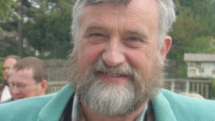 Mand med gråt skæg og hår, der bærer seler og grøn jakke.