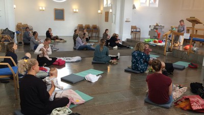 En gruppe af mennesker med børn deltager i en aktivitet i en stor hall med høje vinduer og stoler.