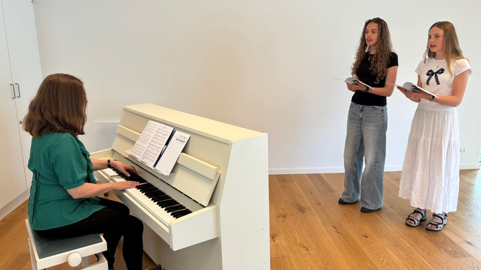 En kvinde spiller piano mens to andre kvinder står ved siden af og læser fra noter.