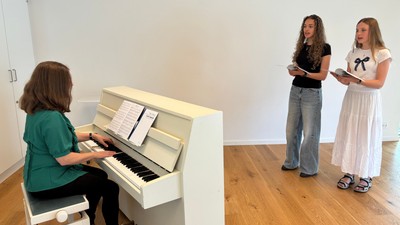 En kvinde spiller piano mens to andre kvinder står ved siden af og læser fra noter.