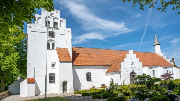 Et hvidt kirkebygning med røde tag og klokketårn, omgivet af grønne træer og en klar blå himmel