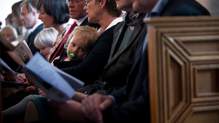 Mennesker, der sidder i kirkebænke, en holder en baby, alle holder bøger eller papirer.