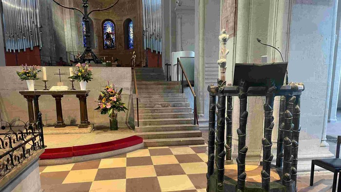 In der Kirche steht ein Pult auf einem Schachbrettboden, umgeben von Blumen und Stufen.