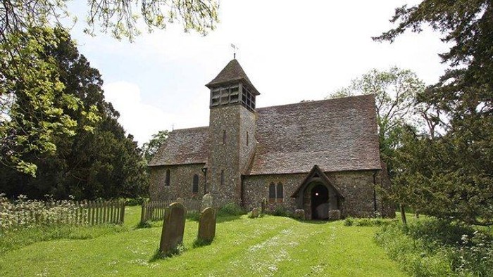Small stone church with a tower and arched entrance, surrounded by a graveyard and trees.