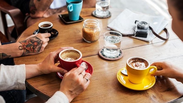 Um einen runden Tisch sitzen Menschen. Auf dem Tisch verschiedenfarbige Kaffee- und Teetassen, Zucker und Wasser und eine Kamera.