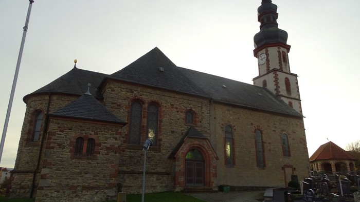 Kirchengebäude mit hohem Turm und Uhr, umgeben von Gräbern und einem Fahnenmast