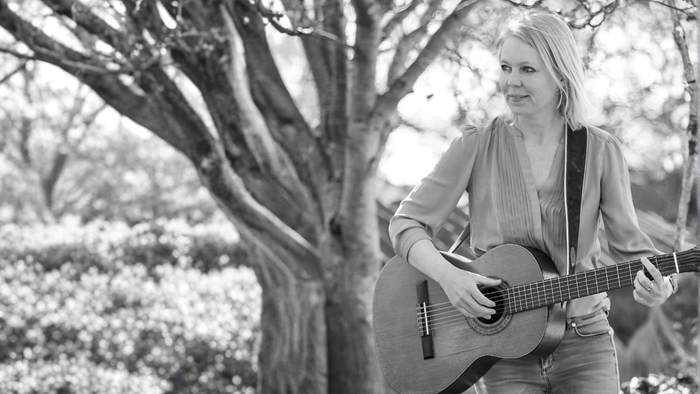 En kvinde spiller akustisk guitar under et stort træ i en fredfyldt udendørs omgivelse.