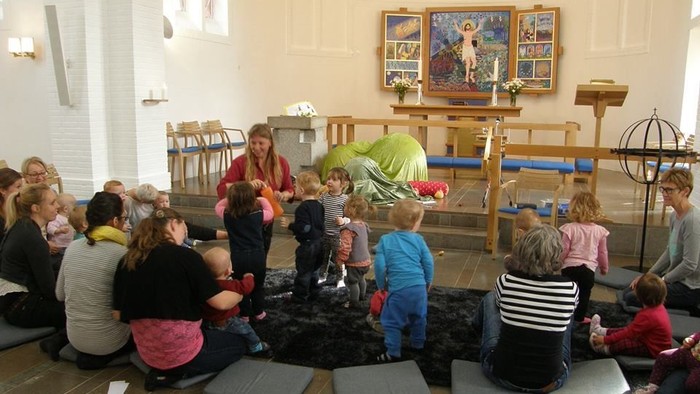 En gruppe børn og voksne sidder i en kirke og lytter til en kvinde, der fortæller en historie.