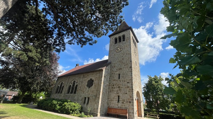 Man seht die Johanneskirche aus Sandstein mit gepflastertem Weg. Das Gebäude hat vorne einen Glockenturm, davor steht eine alte Kirchenbank.
