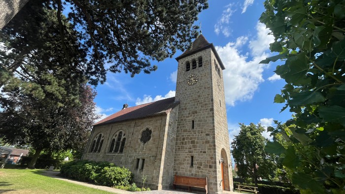 Man seht die Johanneskirche aus Sandstein mit gepflastertem Weg. Das Gebäude hat vorne einen Glockenturm, davor steht eine alte Kirchenbank.