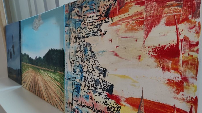 Drei Gemälde auf einem Regal: eines mit einer Landschaft, eines mit abstrakter Kunst und eines mit einer Stadtansicht.