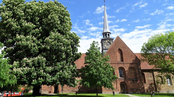 En rød murstenskirke med tårn og klokke, omgivet af grønne træer og en græsplæne