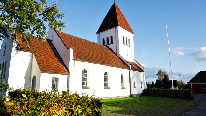 En hvid kirke med røde tag og tårn i en træet og græsområde