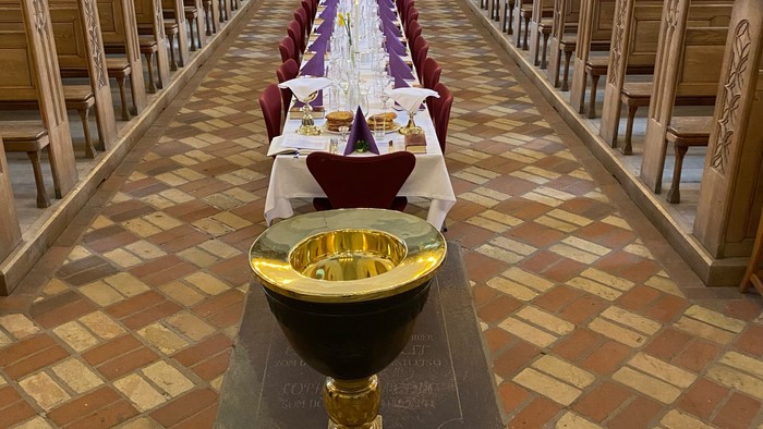 En lang bord med hvidt tæppe og røde stolestøv i en kirke, med en stor guldfarvet kægle på forgrunden