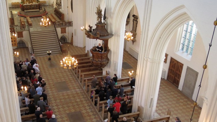 En stor kirke med høje vægge og bjælker, hvor en folkemasse er samlet.