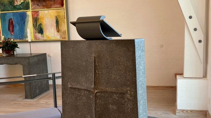 En stor sten med et udhugget kors står i et rum med billeder og trappe.