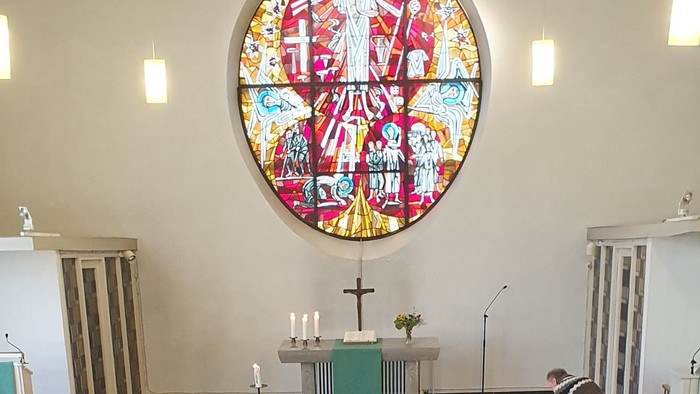 Elegante Kircheninnenausstattung mit einem lebendigen Buntglasfenster über einem schlichten Altar mit Kerzen und Blumenschmuck.
