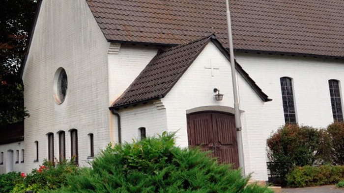 Weiße Kirche mit hohem Turm und braunem Dach, umgeben von Bäumen und Büschen