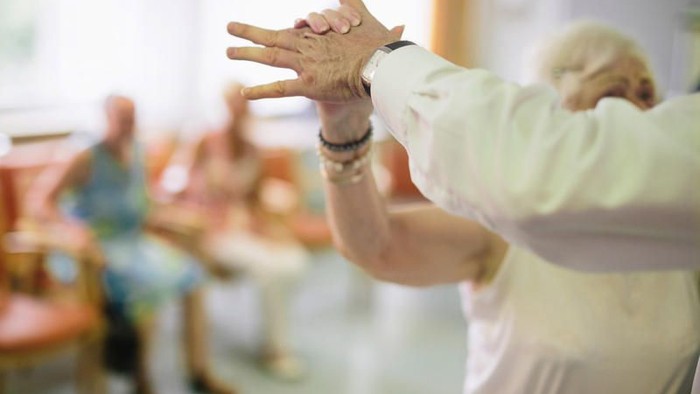 Eine ältere Frau führt eine Handbewegung in einem Raum mit anderen Senioren aus.
