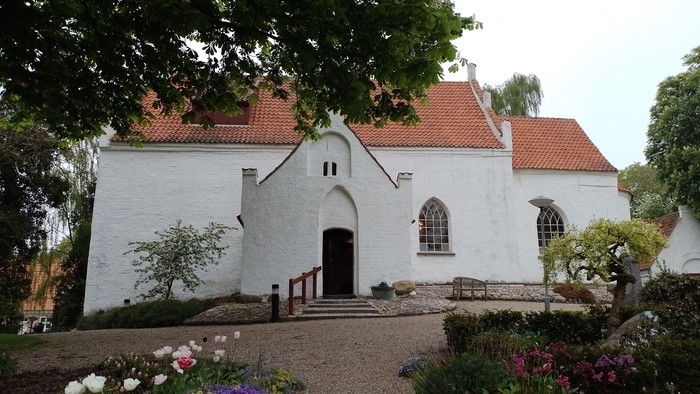 Hvid kirke med rødt tag og kirkegårdens blomster og et grønt træ i forgrunden
