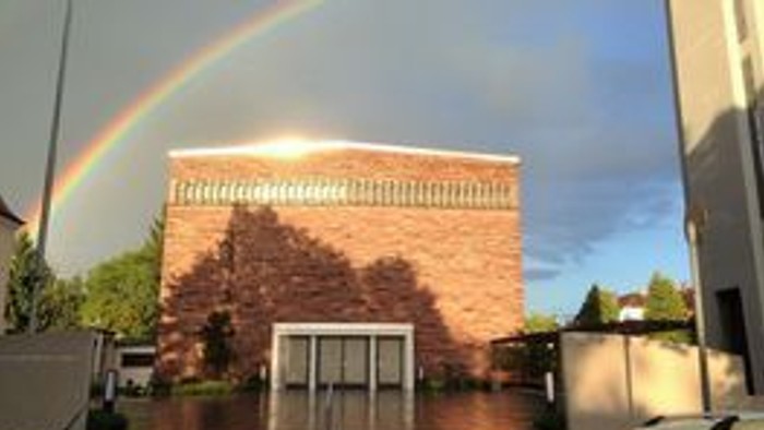 Ein Ziegelgebäude mit einem Regenbogen, der sich am Himmel darüber wölbt, eingerahmt von Bäumen und moderner Architektur.