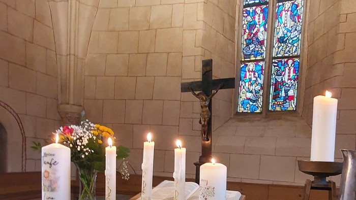 Ein ruhiger Kirchenaltar mit brennenden Kerzen, Blumen und einem Kruzifix hinter bunten Glasfenstern.