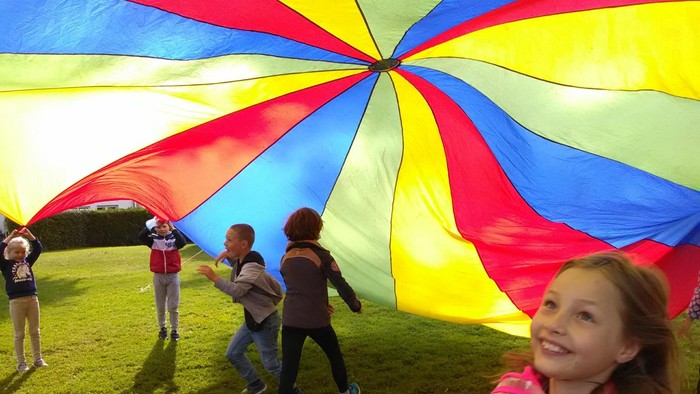 Kinder spielen fröhlich unter einem großen, bunten Fallschirm auf einer offenen Rasenfläche.