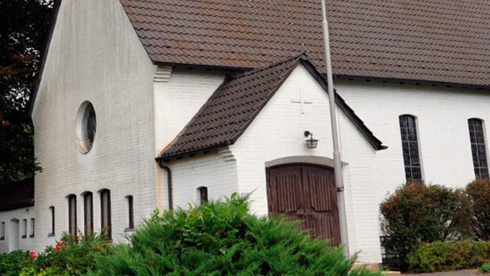 Weiße Kirche mit hohem Turm und braunem Dach, umgeben von Bäumen und Büschen