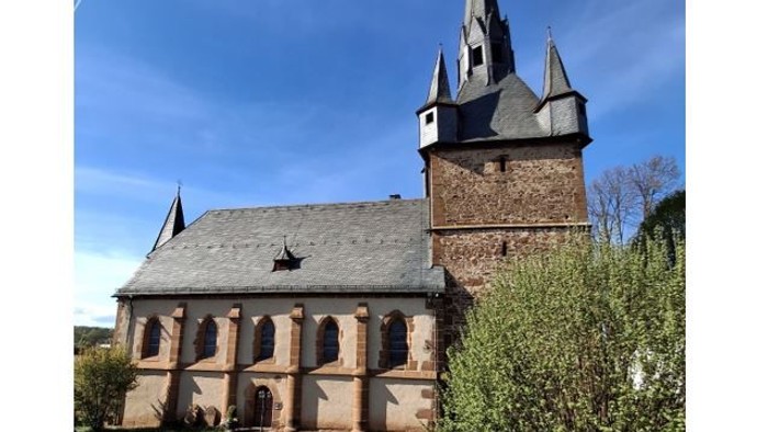 Kirche mit hohem Turmspitz und Rundbogenfenstern, umgeben von grünem Gras und Sträuchern.