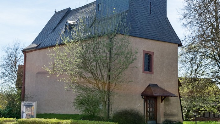 Kirche mit hohem Turm und Kreuz, umgeben von Bäumen und Gras.