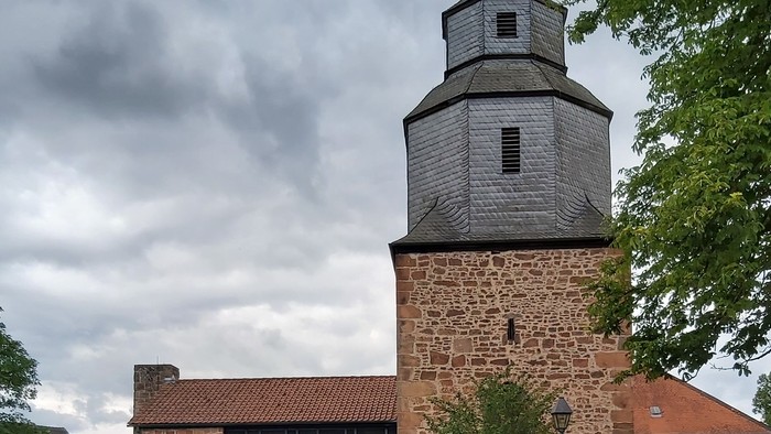 Historische Steinkirche mit modernem Glasanbau, umgeben von Grünflächen und vor bewölktem Himmel gelegen.