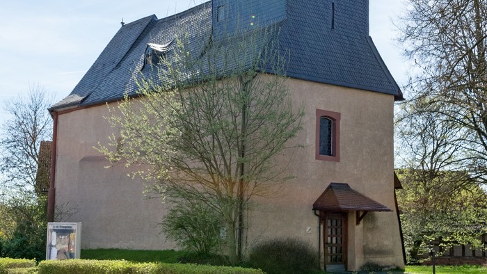 Kirche mit hohem Turm und Kreuz, umgeben von Bäumen und Gras.