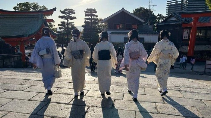 Fem personer i traditionelle japanske kimonoer går sammen i en solbeskinnet tempelgård.
