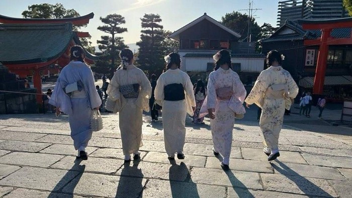 Fem personer i traditionelle japanske kimonoer går sammen i en solbeskinnet tempelgård.