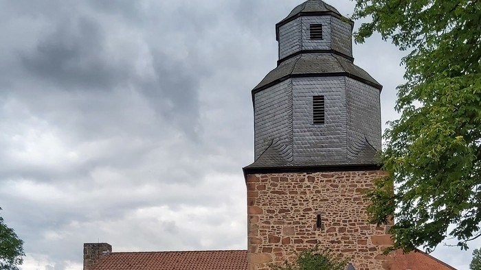 Historische Steinkirche mit modernem Glasanbau, umgeben von Grünflächen und vor bewölktem Himmel gelegen.