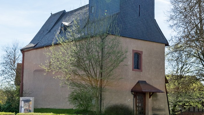 Kirche mit hohem Turm und Kreuz, umgeben von Bäumen und Gras.