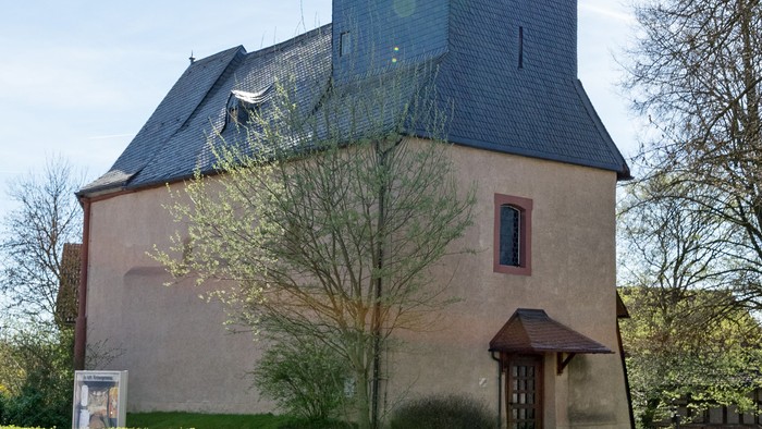 Kirche mit hohem Turm und Kreuz, umgeben von Bäumen und Gras.