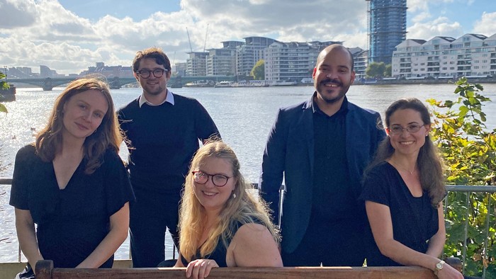 Fem sortklædte sangere poserer ved bænk foran sø