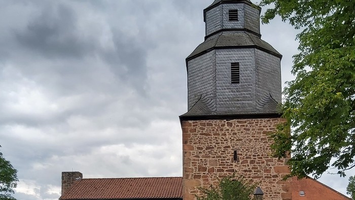 Historische Steinkirche mit modernem Glasanbau, umgeben von Grünflächen und vor bewölktem Himmel gelegen.