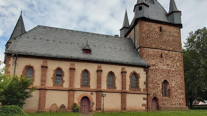 Kirche mit hohem Turmspitz und Rundbogenfenstern, umgeben von grünem Rasen.