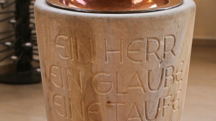 Der Taufstein ist aus Stein gefertigt und hat eine metallene Einfassung.