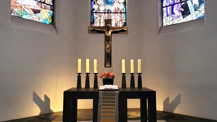 Moderner Kapellenaltar mit Kruzifix, Kerzen und farbenfrohen Buntglasfenstern hinter einem minimalistischen Tisch.
