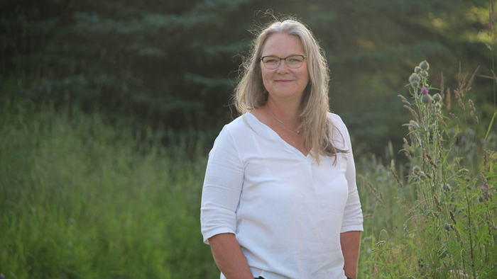En kvinde med blondt hår og briller står på en græsklædt mark.
