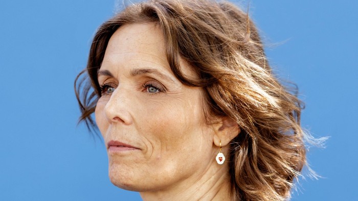 En kvinde med kort brunt hår i en mørk blazer foran en lys blå baggrund.