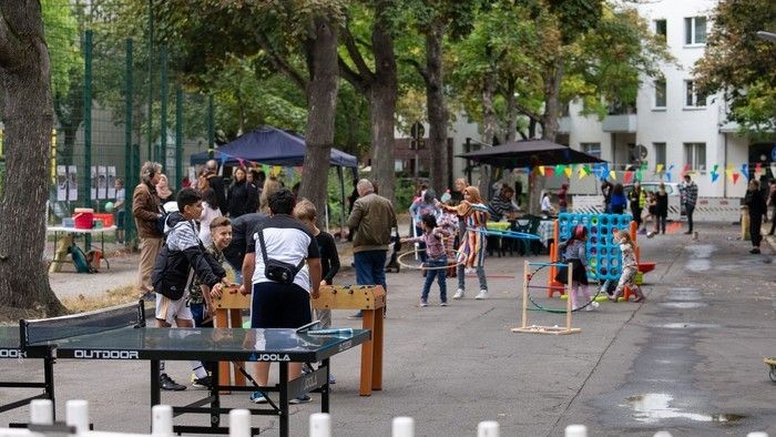 Menschen spielen Tischtennis im Freien, umgeben von Bäumen und Gebäuden.