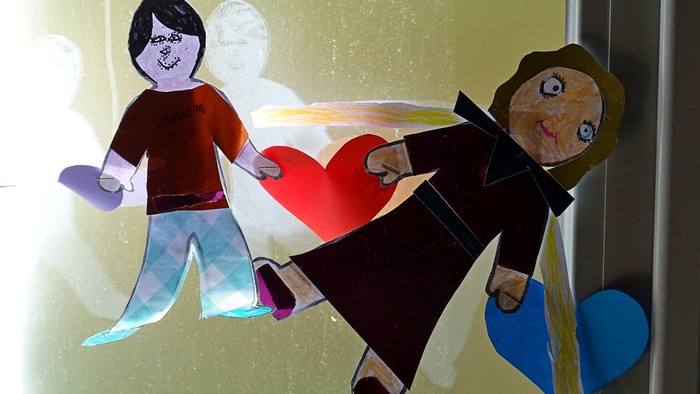 Zwei Papierfiguren halten ein rotes Herz, eine trägt eine blaue Schleife